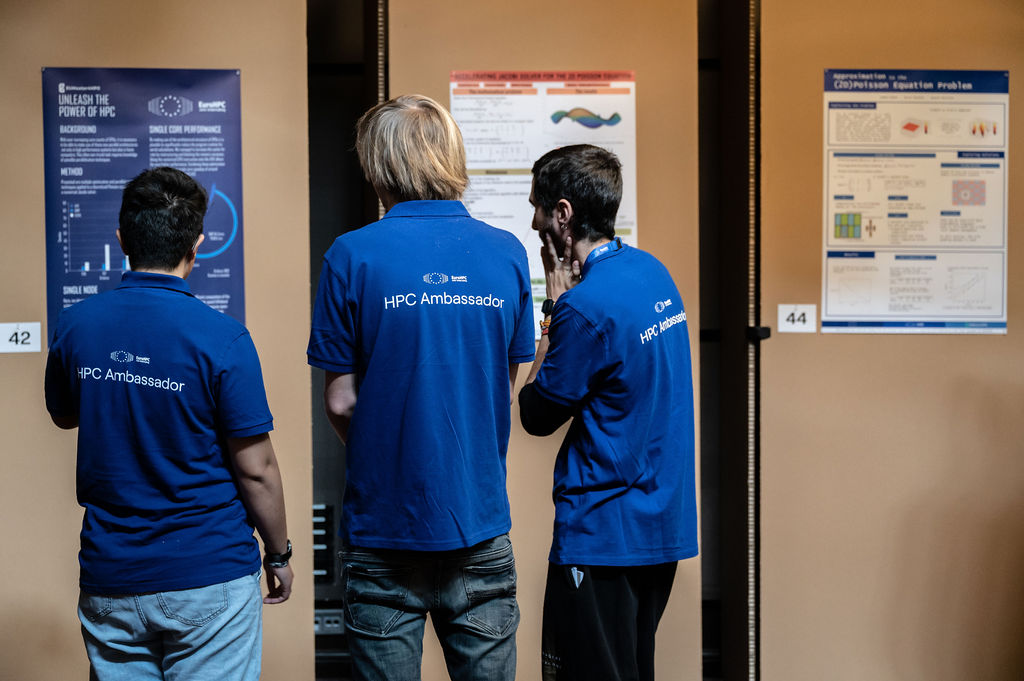Three young people seen from behind, reading a scientific poster. They are wearing blue tops with the words "HPC Ambassador" on the back.
