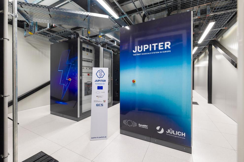 A server room with the JUPITER supercomputer, labeled as Europe’s first exascale system. It’s housed in large cabinets, with a sign showing the logos of the organisations that Jupiter is funded by.